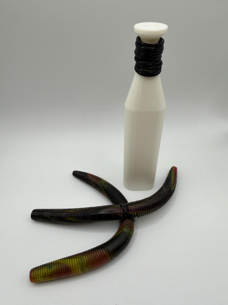 May include: A white bottle with a black ringed neck and a white top. Three dark brown, ribbed, curved fishing lures with red and green accents are arranged in front of the bottle.