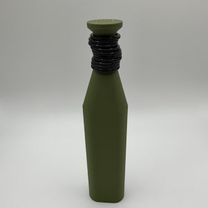 Puede incluir: Una botella verde oliva con cuerpo cil&iacute;ndrico y parte superior c&oacute;nica. La parte superior est&aacute; adornada con una serie de anillos negros, creando un elemento decorativo. La botella est&aacute; sobre un fondo blanco liso.
