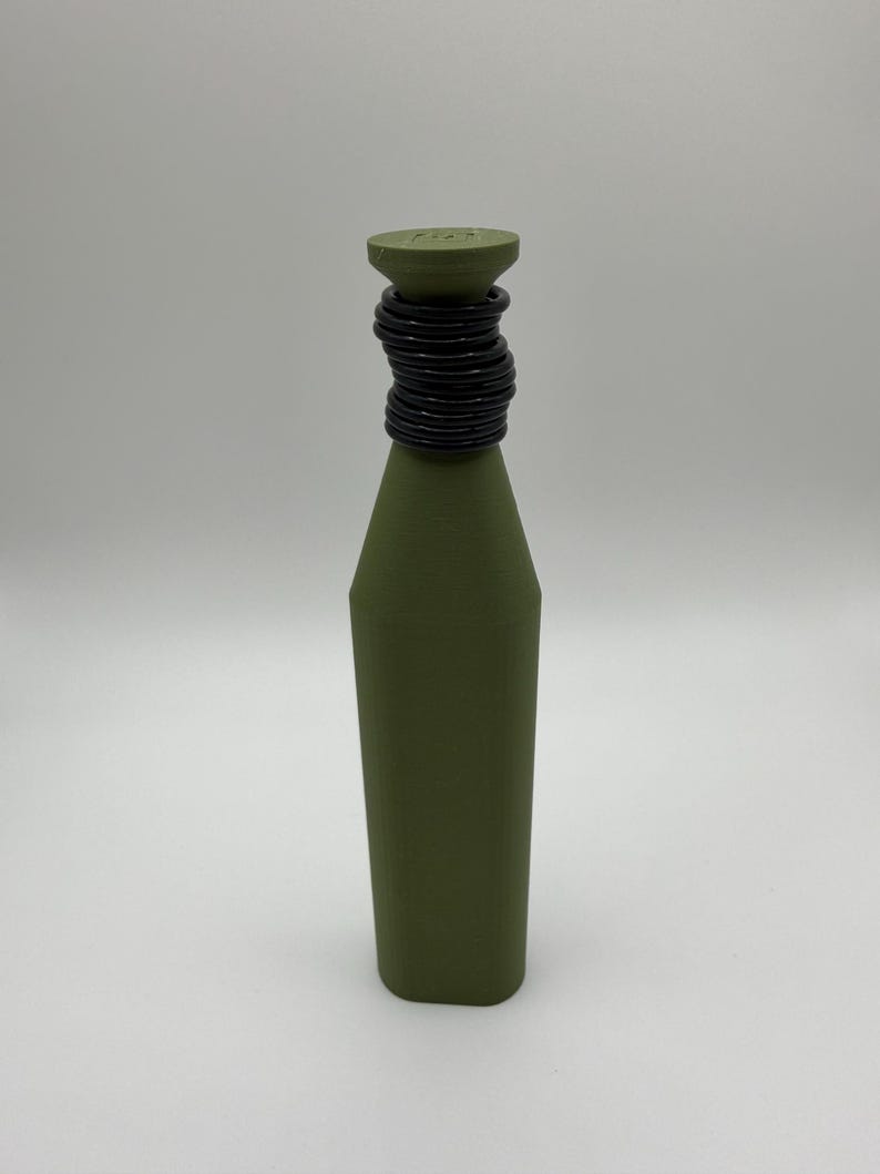 Puede incluir: Una botella verde oliva con cuerpo cil&iacute;ndrico y parte superior c&oacute;nica. La parte superior est&aacute; adornada con una serie de anillos negros, creando un elemento decorativo. La botella est&aacute; sobre un fondo blanco liso.