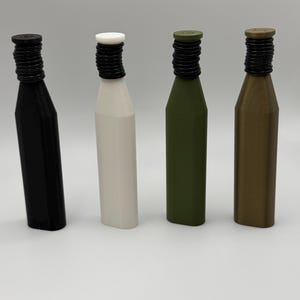 Puede incluir: Cuatro tapones de botella en negro, blanco, verde oliva y bronce. Cada tap&oacute;n tiene un cuello negro estriado y una parte superior plana. Las botellas son cil&iacute;ndricas con una forma ligeramente c&oacute;nica.