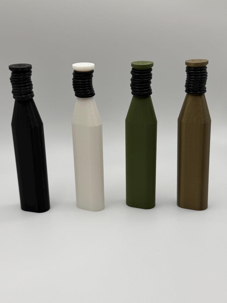 Puede incluir: Cuatro tapones de botella en negro, blanco, verde oliva y bronce. Cada tap&oacute;n tiene un cuello negro estriado y una parte superior plana. Las botellas son cil&iacute;ndricas con una forma ligeramente c&oacute;nica.