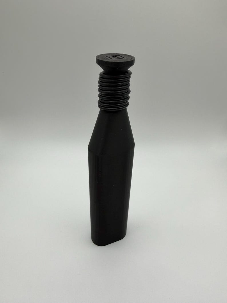 May include: A black bottle stopper with a cylindrical neck and a flat, square top. The neck features a series of concentric rings. The stopper has a geometric, modern design. The bottle stopper is made of a solid, dark material.