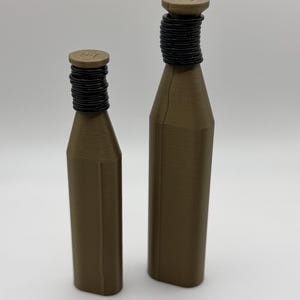 May include: Two brown bottles with black, ridged necks and flat, circular tops. The bottles are different sizes, with the larger one standing taller than the other. The bottles are set against a plain, light background.