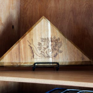 May include: A wooden, triangular tray with a snail and tree design. The tray is displayed on a black metal stand. The tray is made of light brown wood and is displayed on a wooden shelf.