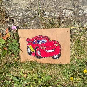 May include: A string art depiction of a red cartoon car with white, yellow, and blue details. The artwork is mounted on brown paper and set on grass, showcasing a handcrafted design.