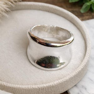 May include: A wide, silver ring with a textured finish. The ring has a curved shape and is presented on a white, circular tray. The background features greenery and a wooden surface.