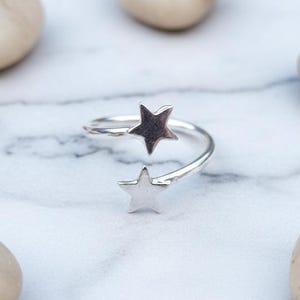 May include: A silver-colored ring with two star-shaped accents. The ring has a wrap-around design, with one star at the top and another at the bottom. The ring is displayed on a white and gray marble surface, surrounded by smooth, light-colored stones.