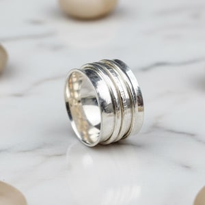 以下が含まれることがあります： マルチバンドデザインのシルバーリング。リングは、積み重ねられた一連のリブ付きバンドを備え、テクスチャード加工の外観を作り出しています。リングは、滑らかなベージュ色の石が散らばる大理石の表面に置かれています。