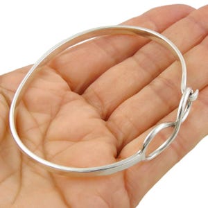 Pode incluir: Uma pulseira de bracelete prateada com um design de símbolo de infinito. A pulseira é feita de metal polido e apresenta um design simples e elegante. A pulseira é mostrada na mão de uma pessoa.
