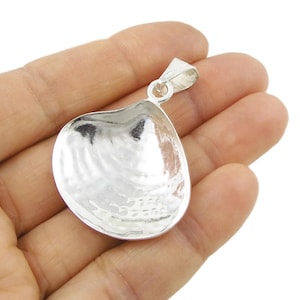 Op de afbeelding: Een zilveren hanger in de vorm van een schelp. De hanger heeft een getextureerd oppervlak en een lus voor een ketting. De hanger wordt in de hand van een persoon gehouden.
