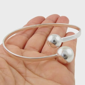 May include: A silver-colored cuff bracelet with two large, round, polished spheres at each end. The bracelet has a simple, open design and is held in a hand.