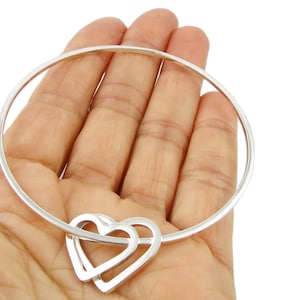 Op de afbeelding: Een zilveren armband met een dubbele hartbedel. De armband wordt in een hand gehouden tegen een witte achtergrond. De armband heeft een eenvoudig, elegant ontwerp, perfect voor dagelijks gebruik.