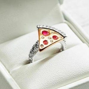 Puede incluir: Un anillo original con forma de porción de pizza, con corteza dorada y «ingredientes» de piedras rojas. La banda plateada está incrustada con pequeñas piedras. Presentado en una caja de joyería blanca.