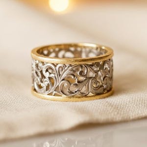 以下が含まれることがあります： 精巧なフィリグリーデザインのツートンカラーリング。リングはゴールドのバンドとシルバーカラーのフィリグリーパターンが特徴です。フィリグリーデザインは、繰り返しの花のパターンです。リングはニュートラルカラーの布の上に置かれています。