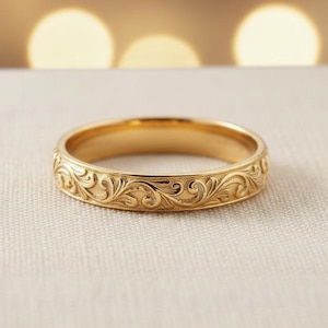 以下が含まれることがあります： 彫刻された花柄のゴールドリング。リングは、背景にぼやけたボケ効果のある、柔らかくニュートラルな背景に配置されています。