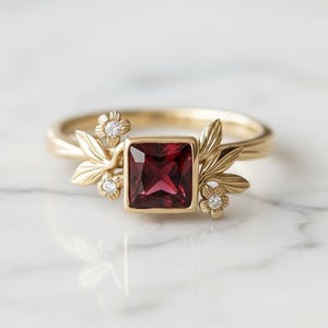 Peut inclure: Une bague en or ornée d'une pierre rouge foncé carrée. La bague est ornée de motifs floraux et de feuilles, ainsi que de petits diamants transparents. La bague est posée sur une surface en marbre blanc.