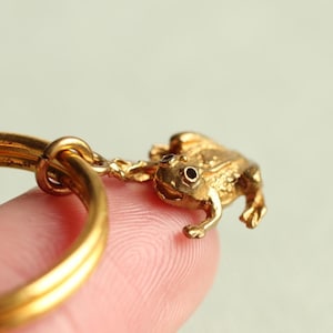 May include: A gold-coloured keyring with a detailed frog charm. The frog is attached to a circular ring, creating a whimsical accessory. The keyring is made of a shiny, metallic material.