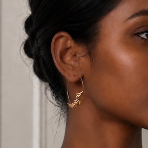 May include: A gold hoop earring featuring a delicate, textured leaf or branch motif attached to the lower curve of the hoop. The metal has a polished, reflective finish, and the design follows the circular shape of the earring wire.