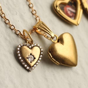 May include: A gold chain necklace featuring two heart-shaped pendants. One pendant is a smooth, matte gold locket. The second pendant is a smaller heart with a central clear stone surrounded by a border of tiny pavé-set stones on a gold base.