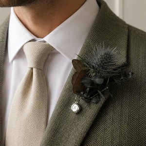 Op de afbeelding: Een corsage met een donkerblauwe distel en gedroogd blad aan een groene blazer met visgraatmotief. Een kleine, ronde zilveren bedel met de letter H hangt aan de basis van het bloemstuk, gedragen over een wit overhemd en een beige das met visgraatmotief.