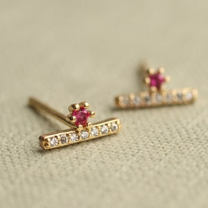 May include: A pair of gold-coloured stud earrings. Each earring has a small, bright red gemstone at the top and a horizontal bar with several small, clear gemstones. The earrings are set against a neutral background.