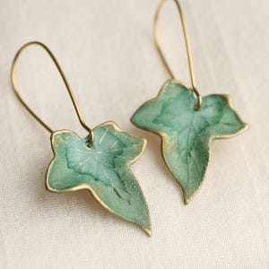 May include: A pair of leaf-shaped earrings. Each earring has a green leaf design with gold-coloured accents. The earrings have a long, curved gold-coloured wire for the ear. The leaves have a textured appearance.