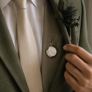 Puede incluir: Un colgante de foto redondo en tono plateado prendido en un chaleco beige, usado bajo un blazer verde oliva de espiga con una camisa blanca de cuello y una corbata beige de espiga. Un boutonniere de cardo oscuro está sujeto a la solapa.