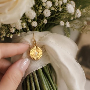 May include: A circular gold-toned pendant featuring a sunburst pattern with a small central white stone, suspended from a thin metal hook and attached to a white ribbon wrapped around a bouquet of white roses and baby's breath flowers.
