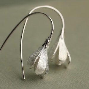 Op de afbeelding: Een paar zilveren oorbellen met een textuurrijke, matte afwerking. Elke oorbel heeft de vorm van een delicate, naar beneden gerichte bloemknop met een gebogen draadhaak die naar boven uitsteekt, wat zorgt voor een sierlijk en langwerpig silhouet.