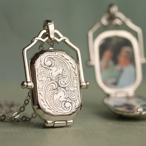 May include: A silver-toned metal locket pendant on a chain. The front of the locket features an intricate engraved scrollwork pattern. A second locket is positioned behind it, shown in an open state, revealing two small photographs inside the frame.
