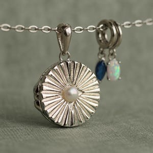 May include: A silver-toned circular locket pendant featuring a sunburst pattern with a central white pearl. The locket hangs from a silver chain alongside two small charms, one with a dark blue stone and the other with an iridescent white stone.