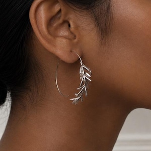 May include: A silver hoop earring featuring a delicate, textured leaf or branch motif along the lower curve. The earring is constructed from a thin, polished metal wire that forms a circular shape, with the botanical design attached to the bottom.