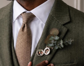 May include: A white collared shirt worn with a tan herringbone tie and a dark olive green herringbone blazer. A silver circular locket containing two small photographs is pinned to the lapel alongside a sprig of green eucalyptus leaves.