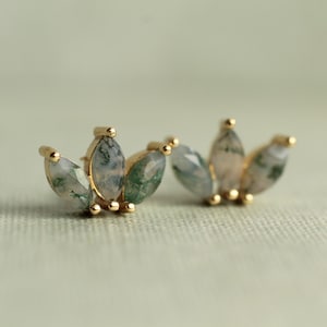 May include: Two gold-coloured stud earrings are shown. Each earring has three marquise-cut gemstones in green and white, set in gold-coloured metal. The earrings have a delicate, nature-inspired design, suitable for everyday wear.