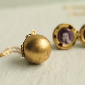 Puede incluir: Un collar con un relicario esférico dorado de estilo vintage. El relicario se abre para revelar una pequeña fotografía. La cadena es delicada y dorada. El relicario tiene una apariencia antigua y desgastada.