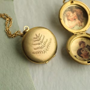 May include: A round gold-toned metal locket pendant on a matching chain. The front of the closed locket features an engraved fern leaf design. An open locket beside it reveals two circular interior compartments, each containing a vintage-style photograph.