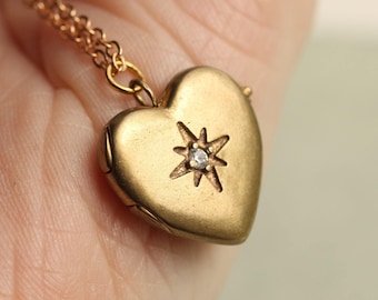 May include: A gold-toned heart-shaped locket pendant suspended from a matching gold-toned chain. The locket features a central starburst engraving with a small, clear, round-cut stone set in the middle of the star design on the front surface.