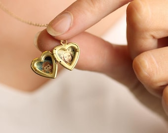 May include: A gold-toned heart-shaped locket pendant suspended from a delicate chain. The locket is open, revealing two interior compartments, each containing a small, circular photograph of a person's face. The metal frame is smooth and polished.