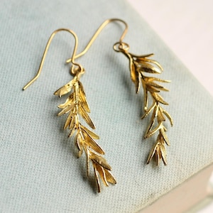 May include: A pair of gold-colored earrings. Each earring features a delicate, leaf-like design with intricate detailing. The earrings hang from simple, curved ear wires. The background is a soft, muted blue.