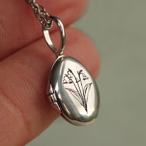 May include: An oval silver locket pendant suspended from a matching silver chain. The front surface of the polished metal locket features a detailed black line drawing of a lily of the valley flower with leaves and multiple small bell-shaped blossoms.