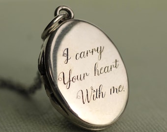 Könnte beinhalten: Ein ovaler silberner Medaillon-Anhänger mit einer polierten metallischen Oberfläche und dem eingravierten kursiven Text I carry Your heart With me. Das Medaillon verfügt über eine kleine kreisförmige Öse an der Oberseite zur Befestigung.