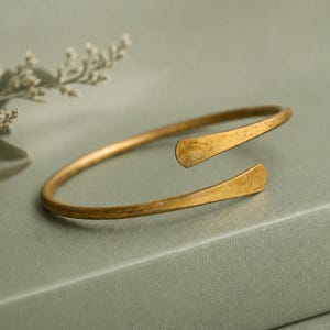 May include: A gold-toned metal cuff bracelet with a smooth, open-ended design. The bracelet features a slightly hammered, matte finish and rounded, tapered ends. The metal has a warm, muted yellow hue with subtle variations in texture and light reflection.