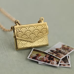 May include: A gold-toned metal envelope locket pendant on a matching chain, featuring an embossed fan-shaped pattern on the front flap. Several small, rectangular photographic prints showing people are arranged near the pendant.