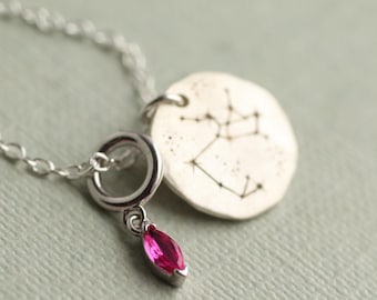 Puede incluir: Un collar de plata con un colgante que muestra una constelación y una pequeña gema rosa. El colgante redondo tiene una superficie texturizada. La gema es de color rosa vibrante y tiene forma de marquesa.