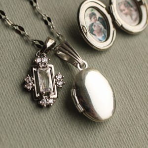 May include: A silver-toned necklace featuring a rectangular pendant with a central baguette-cut clear stone surrounded by small round stones, alongside an oval locket that is open to reveal two small circular photographs of people.