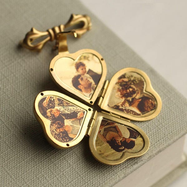 Pode incluir: Um relicário de metal dourado em forma de trevo de quatro folhas com um gancho superior em forma de laço. O relicário está aberto, revelando quatro compartimentos em forma de coração, cada um contendo uma pequena fotografia em tom sépia.