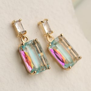 May include: A pair of gold-coloured earrings with rectangular iridescent crystals. Each earring has a small, clear rectangular stone at the top. The larger crystals display a rainbow of colours, including pink, blue, and yellow.