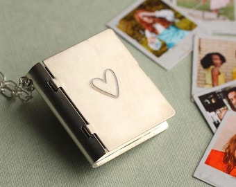 May include: A gold-toned metal locket shaped like a small book with a heart engraved on the front cover. The locket is attached to a silver-colored metal chain link necklace. The surface of the locket has a brushed finish and a hinged spine.