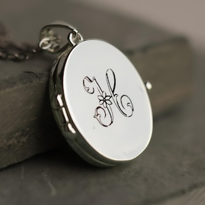 May include: A round silver locket pendant featuring a polished metallic surface with a dark engraved letter K in a decorative script font. The locket includes a small integrated bail at the top for attachment to a chain, showcasing a smooth finish.