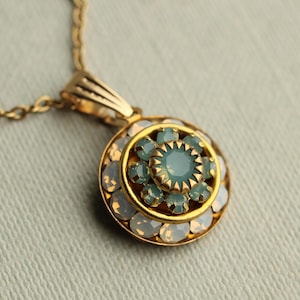 May include: A gold-toned pendant necklace featuring a circular design. The pendant has a central light blue gemstone surrounded by smaller light blue and white gemstones. The necklace chain is also gold-toned.